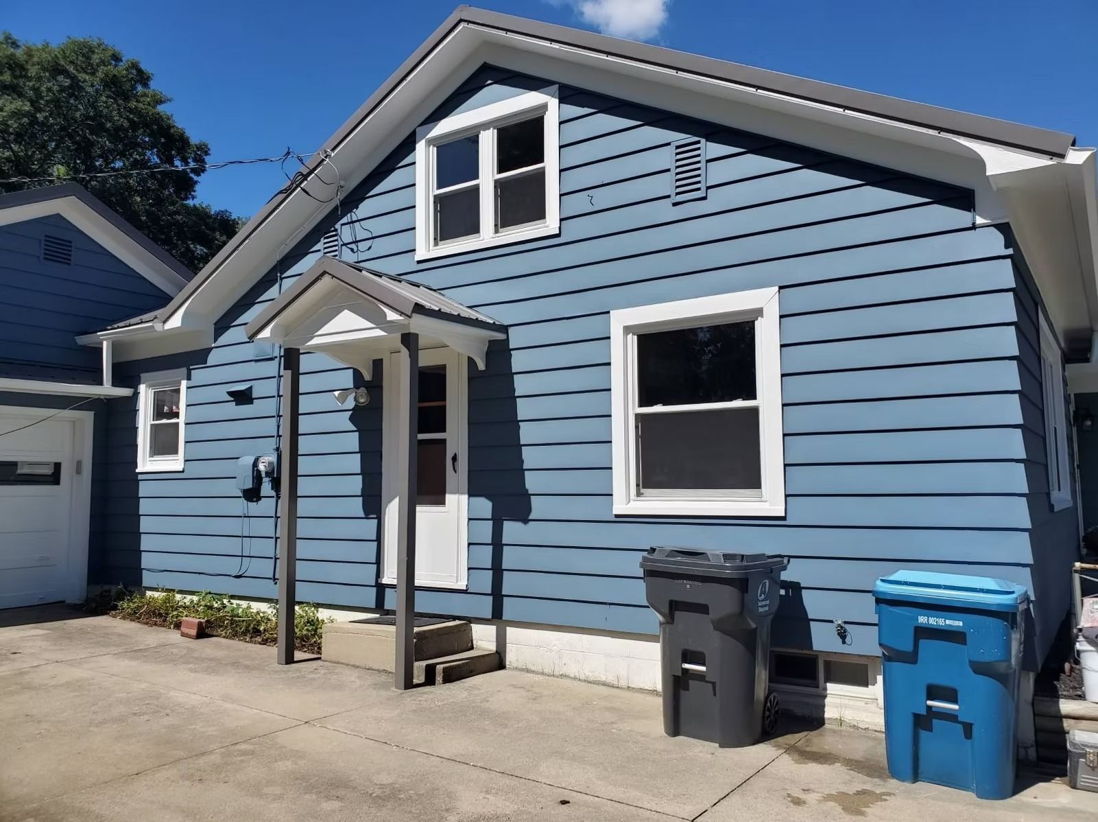 A blue, one-story house with a small front porch, white trim, and two trash cans on a concrete driveway.