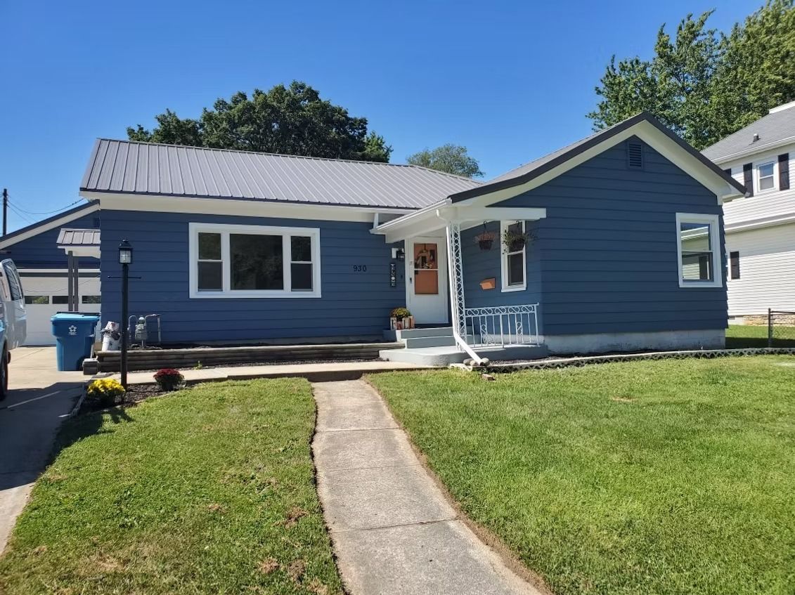 A single-story blue house with a gray metal roof, a white front door, and a concrete walkway leading to the entrance.