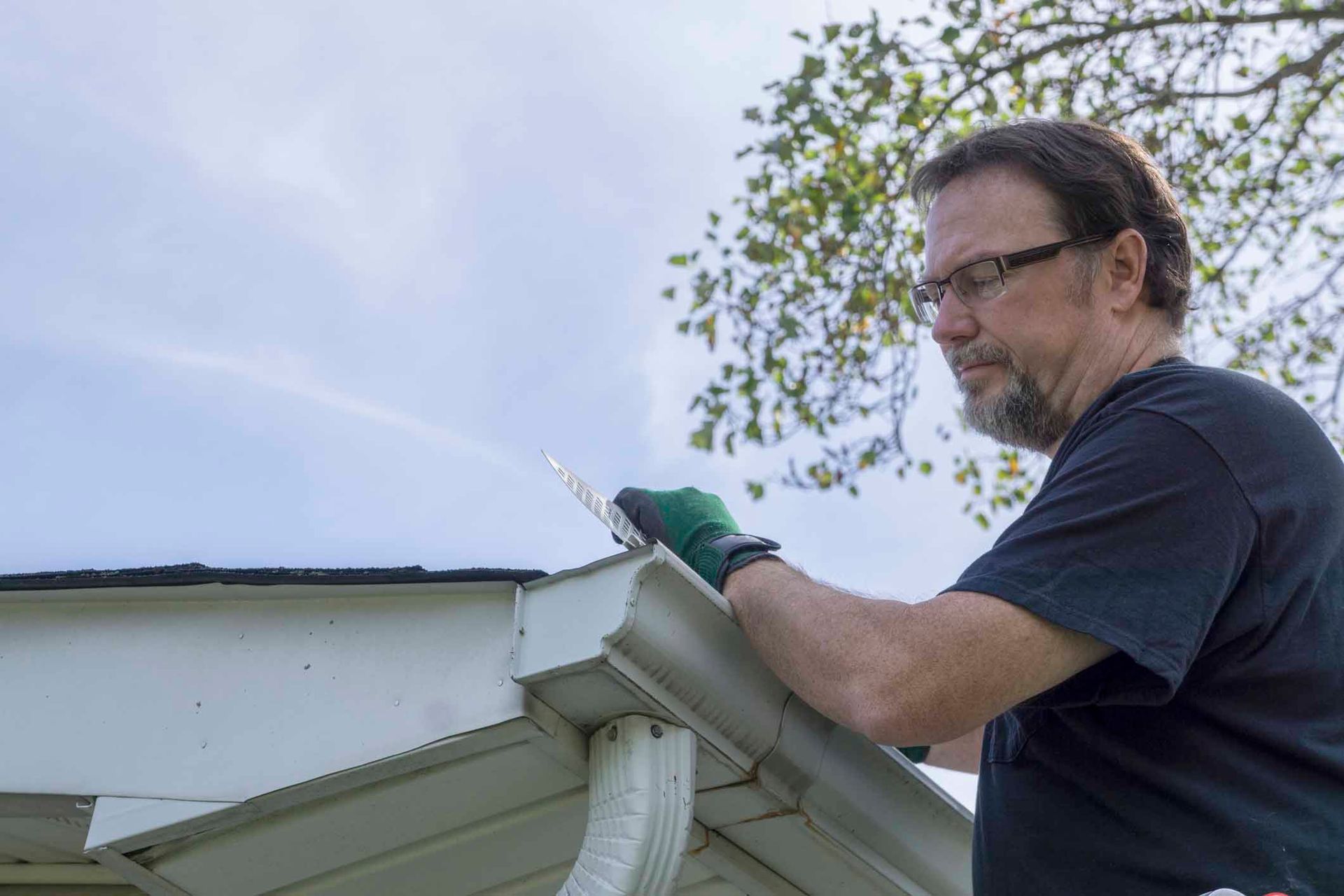 Person cleaning roof gutter with gloved hands on ladder — home maintenance and repair outdoors.
