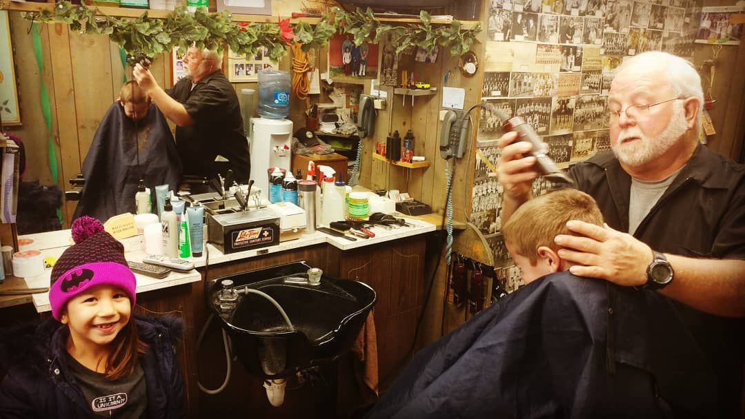 Hair salon with stylist cutting a client’s hair; smiling child in pink hat in foreground.