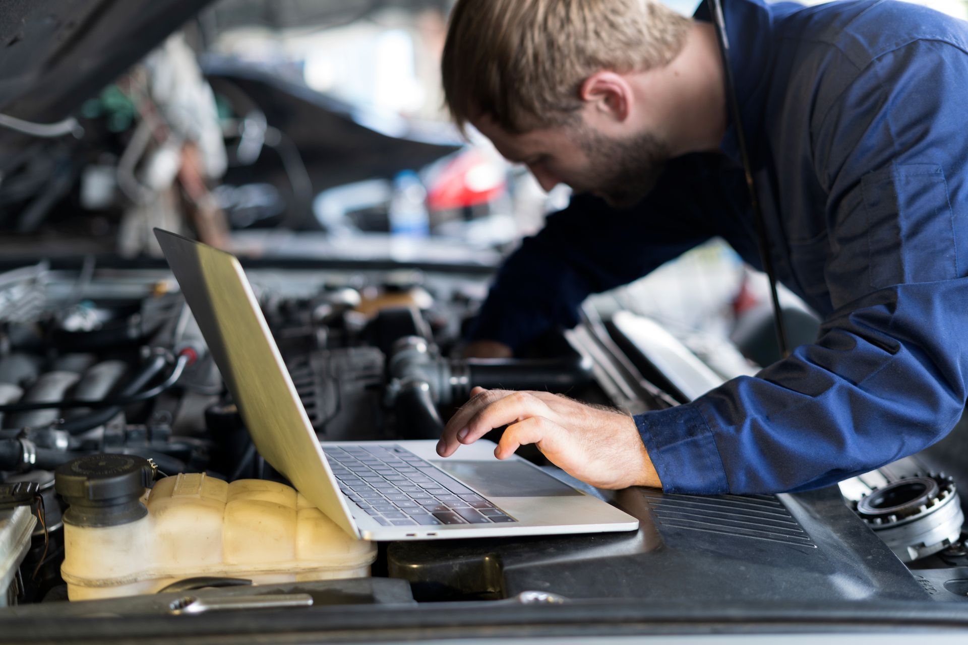 A man is working on a car engine while using a laptop computer.