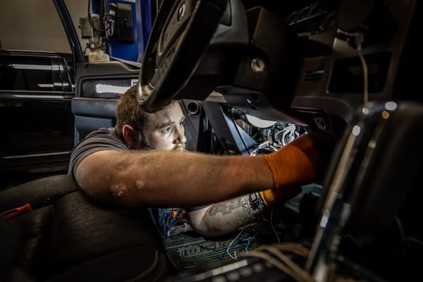 Mechanic working under a car dashboard in a dim garage, focused on wiring.