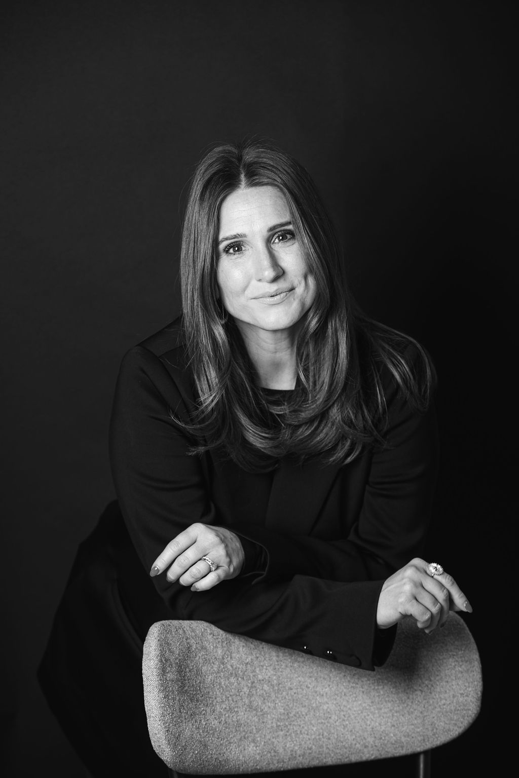 Woman with long hair in a dark blazer, leaning on a textured chair against a black background.