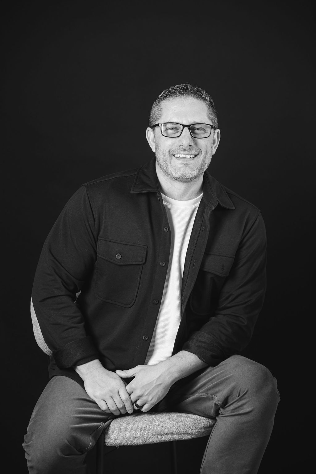 Smiling person seated on a stool against a dark background, wearing glasses, a black jacket, and light jeans