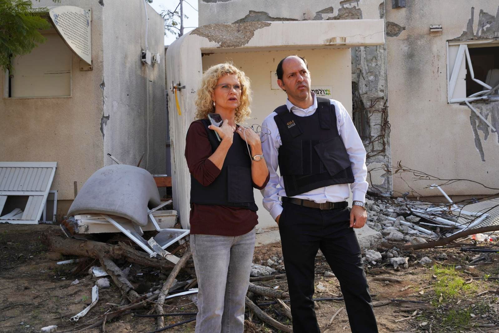 Two people stand amid rubble outside a damaged building, looking concerned.