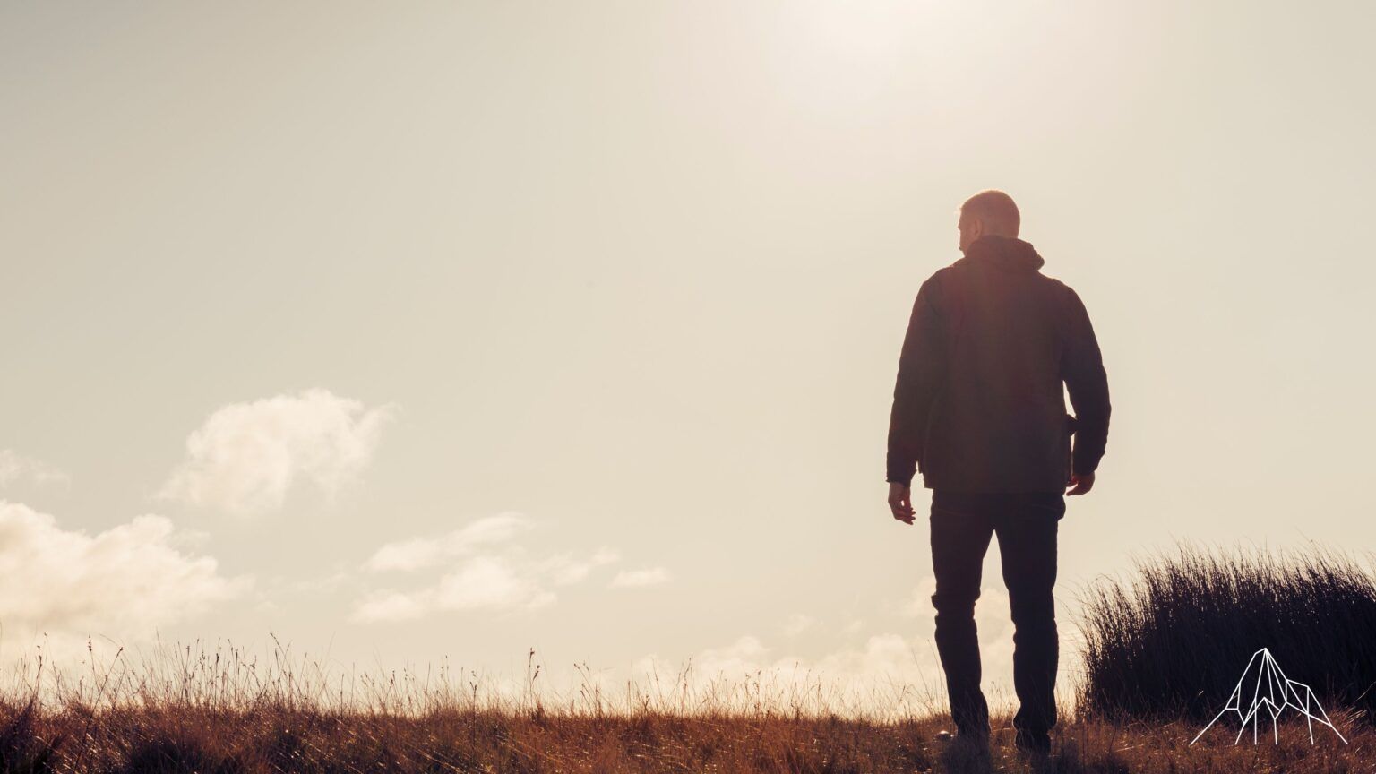 Silhouetted person standing in a grassy field under a bright, hazy sky at sunset