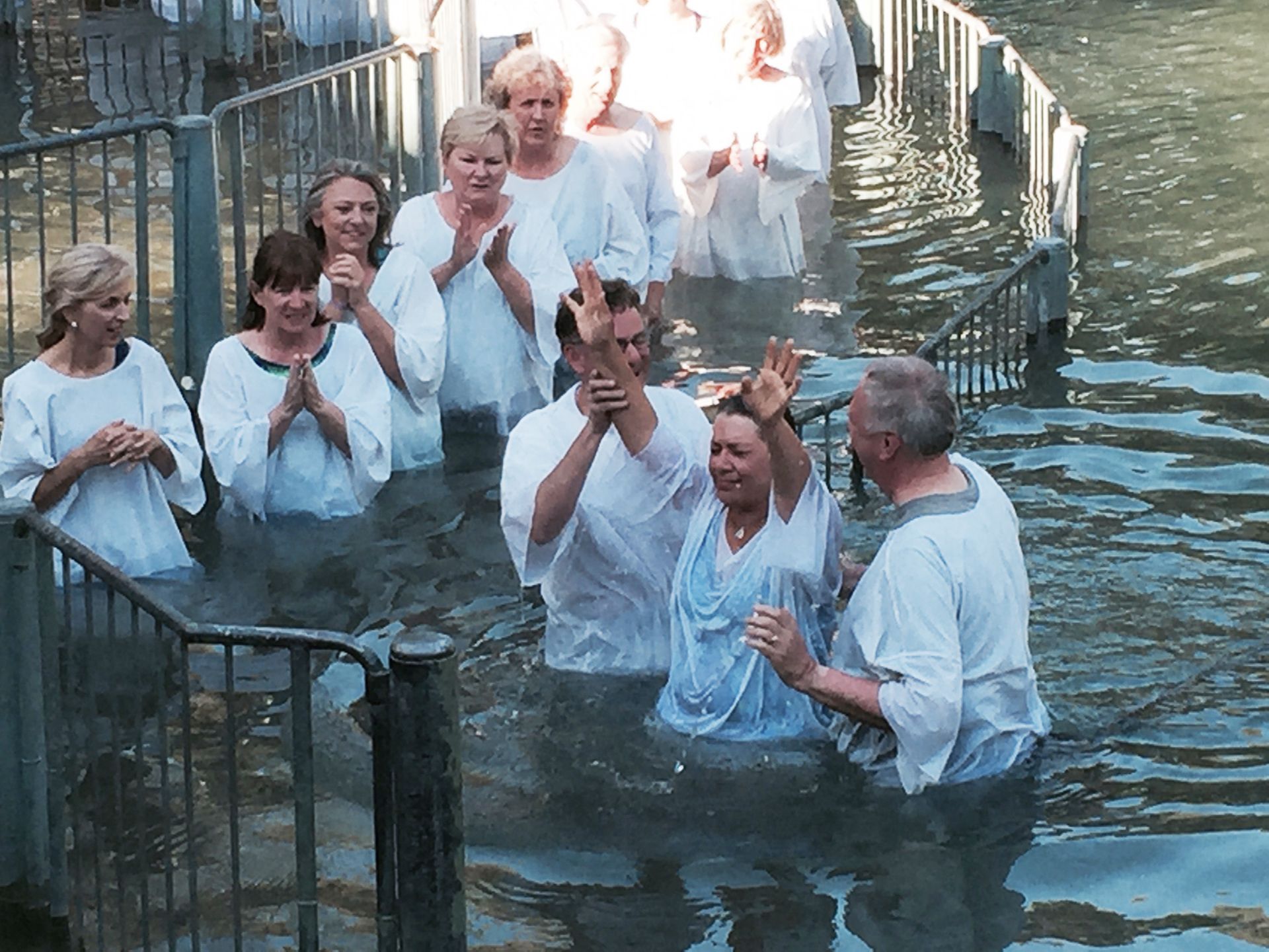 People standing in a shallow water baptism pool, some smiling and clapping in white shirts