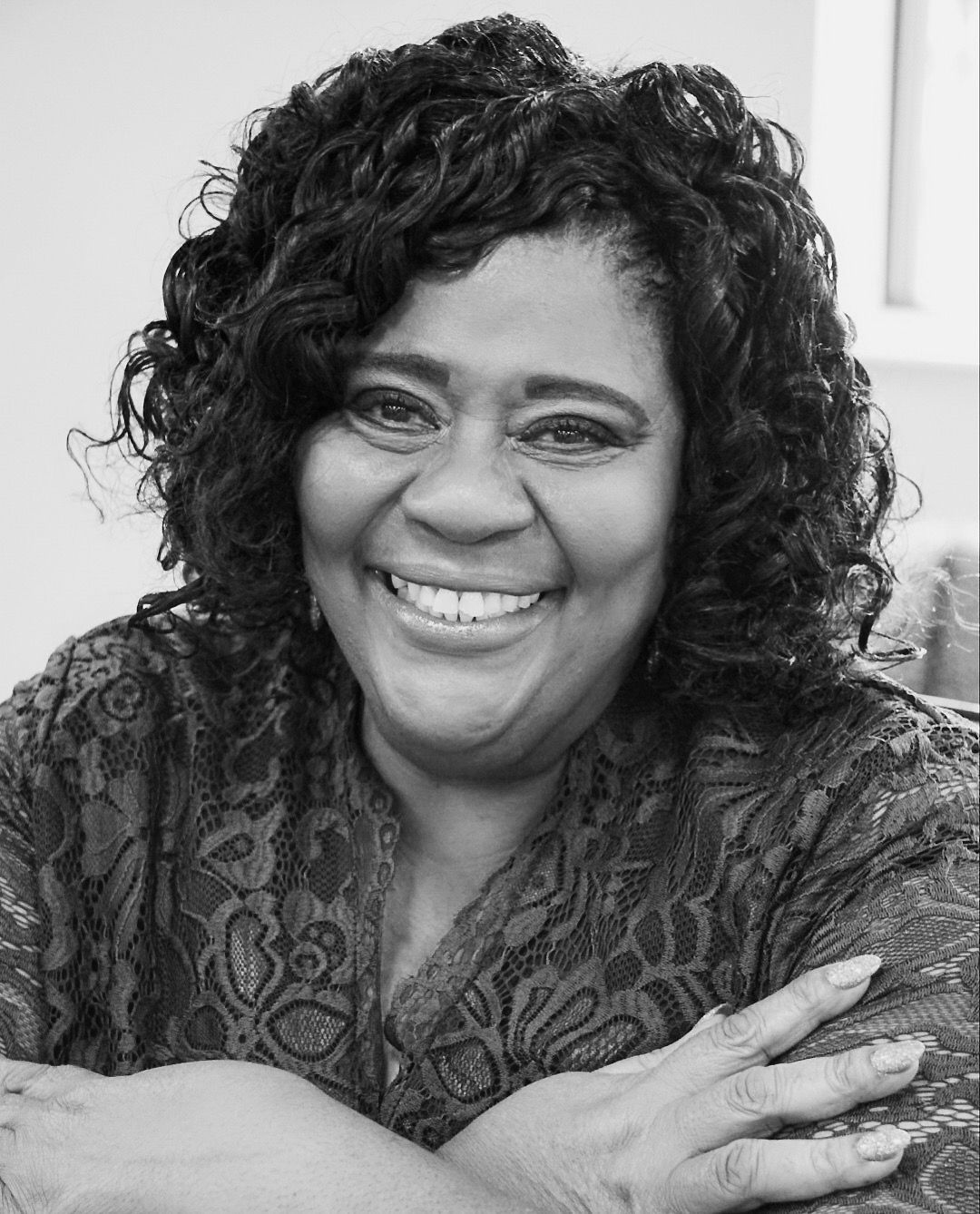 Smiling woman with curly hair and patterned top, arms crossed, in a bright indoor setting