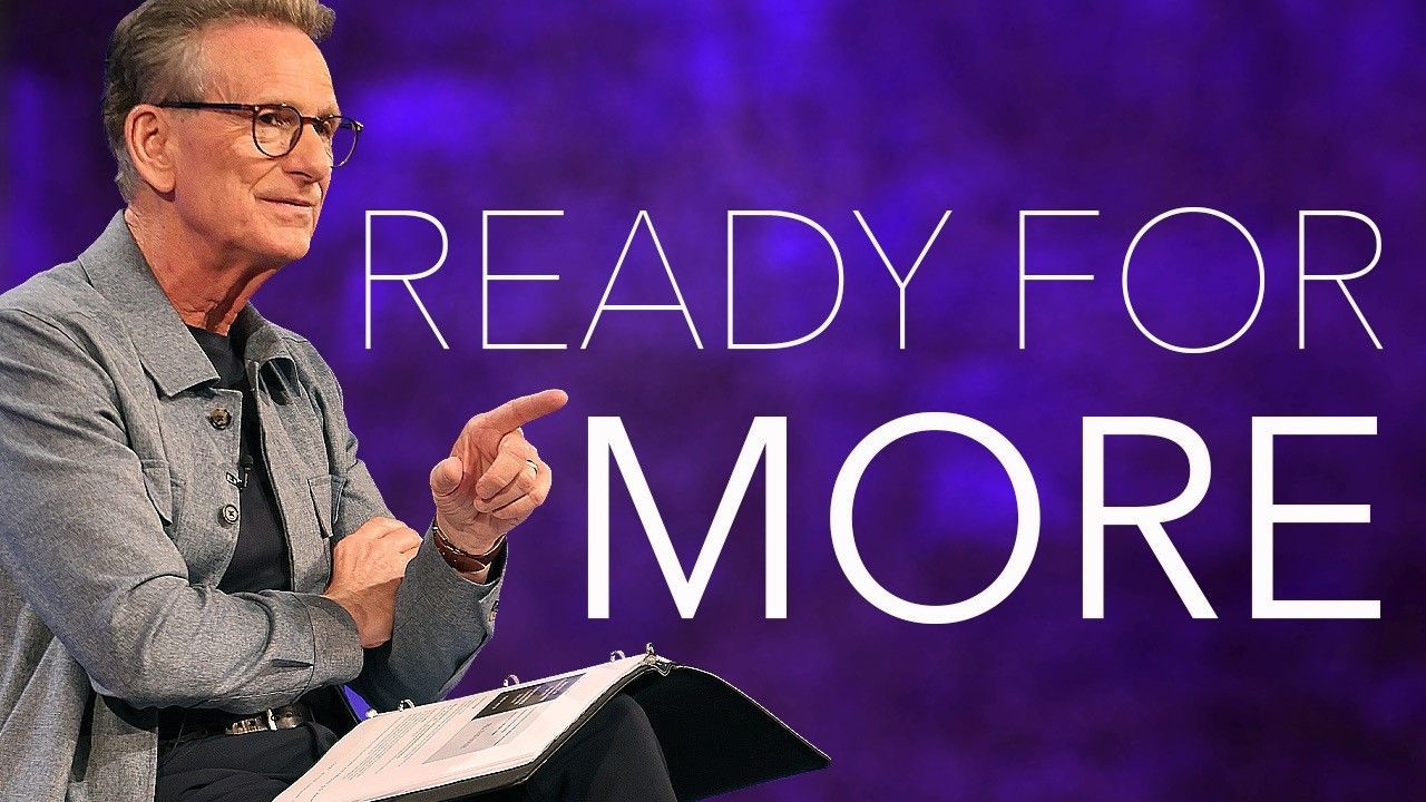 Man onstage in a gray shirt gesturing beside the words “READY FOR MORE” on a purple background