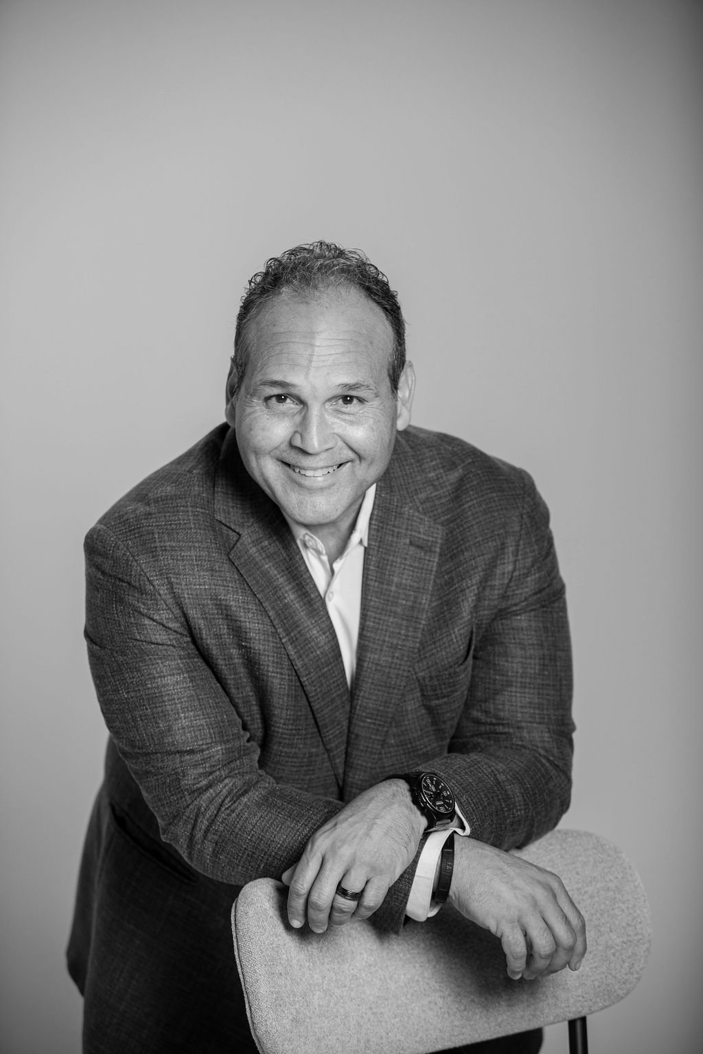 Smiling man in a blazer leaning on a chair, against a plain gray background.