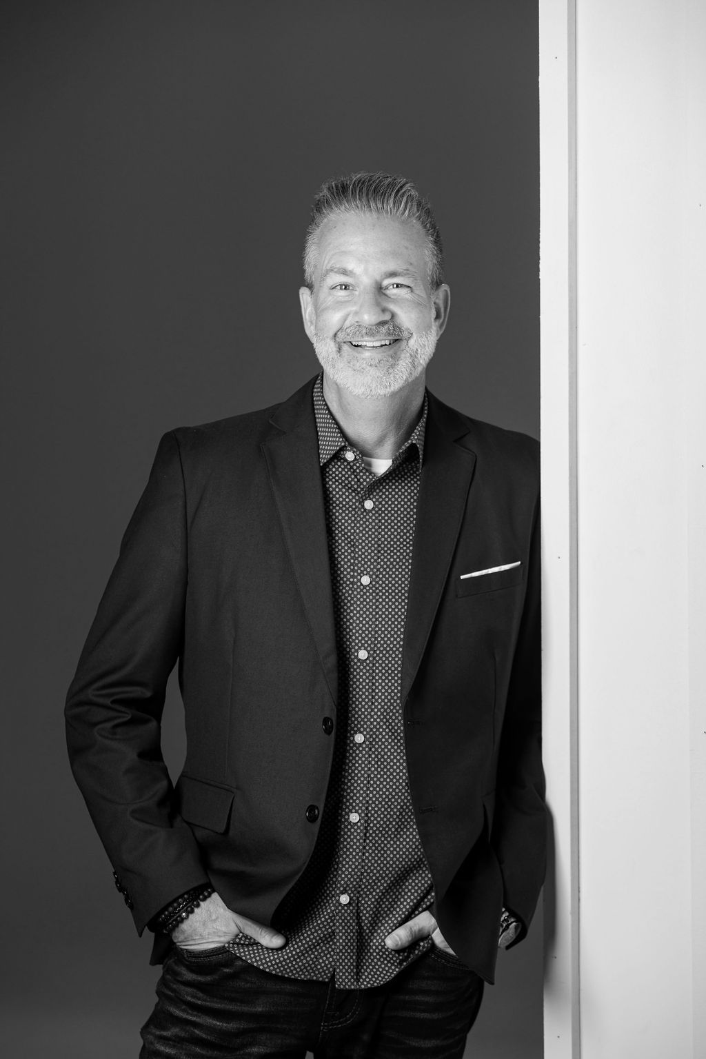 Smiling man in a dark blazer stands against a plain wall, hands in pockets.