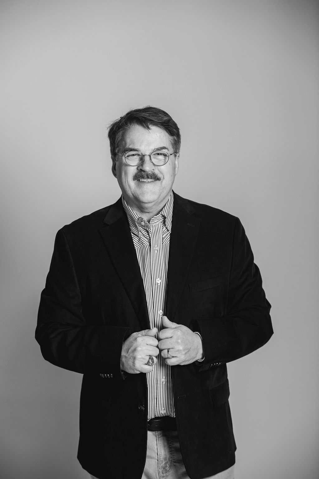 Black-and-white portrait of a man in a dark blazer and striped shirt, standing against a plain background.