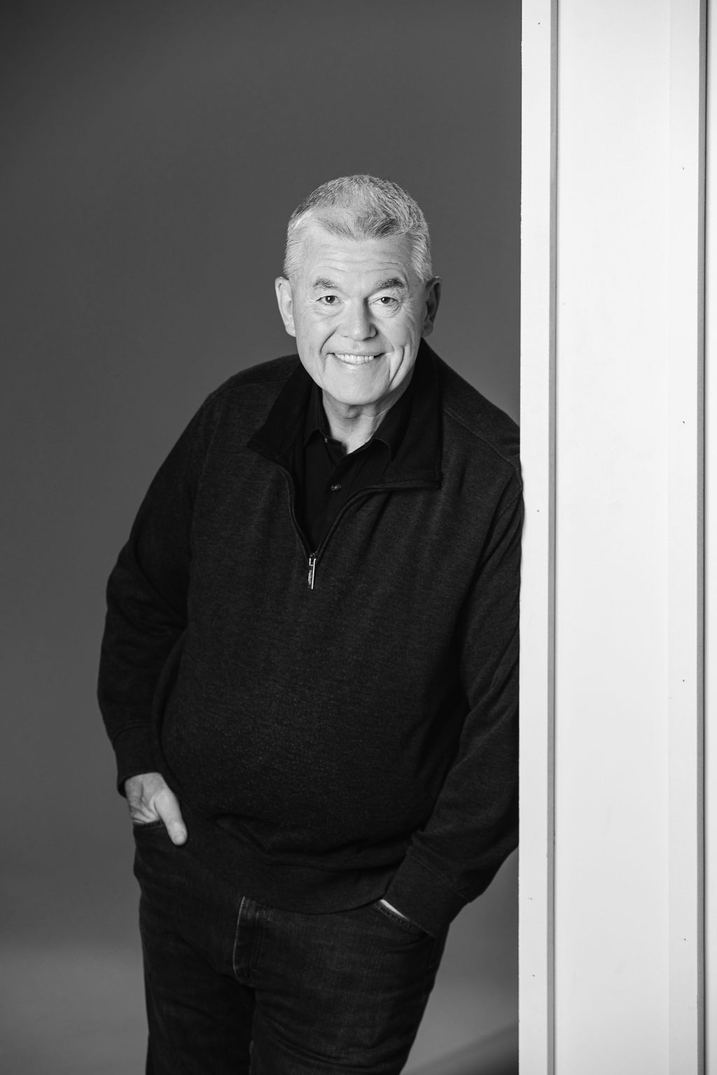 Man in a dark sweater leaning against a wall, smiling at the camera.