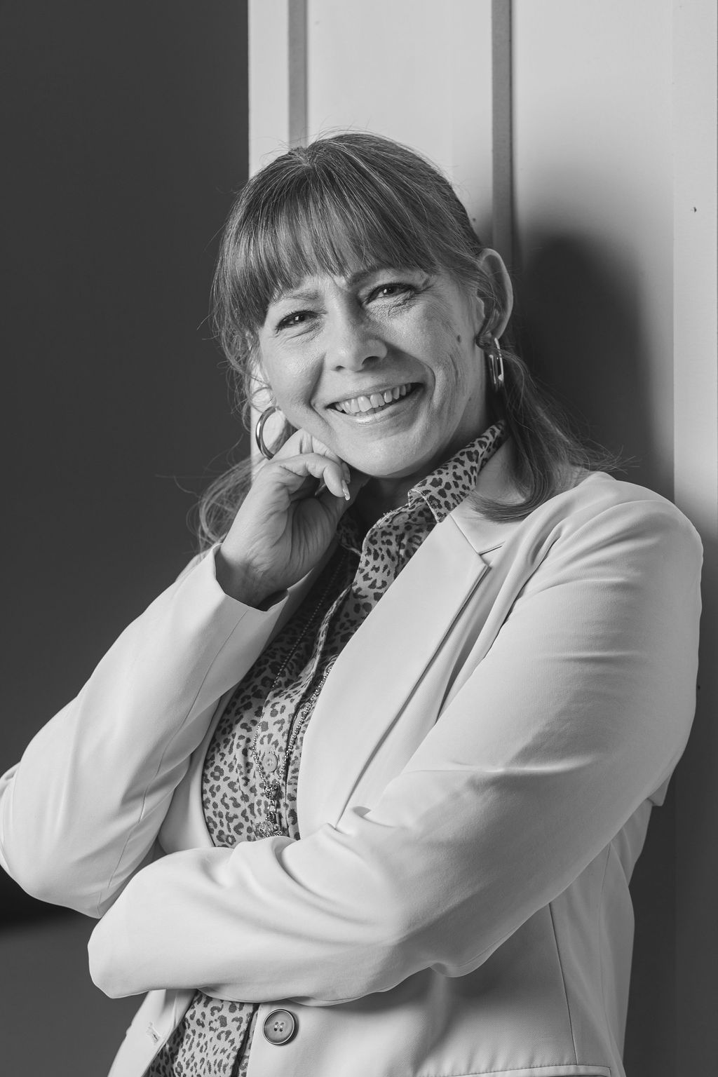 Smiling woman in a light blazer leaning against a wall, resting her hand on her face.