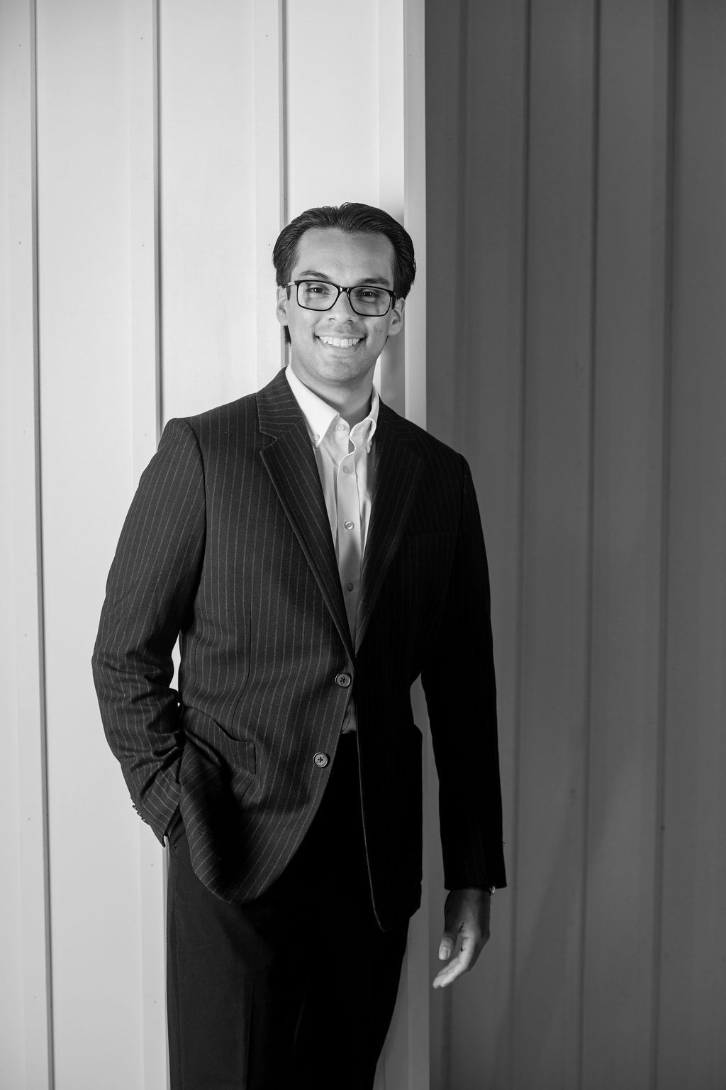 Man in a dark pinstripe suit and glasses stands smiling against a paneled wall.