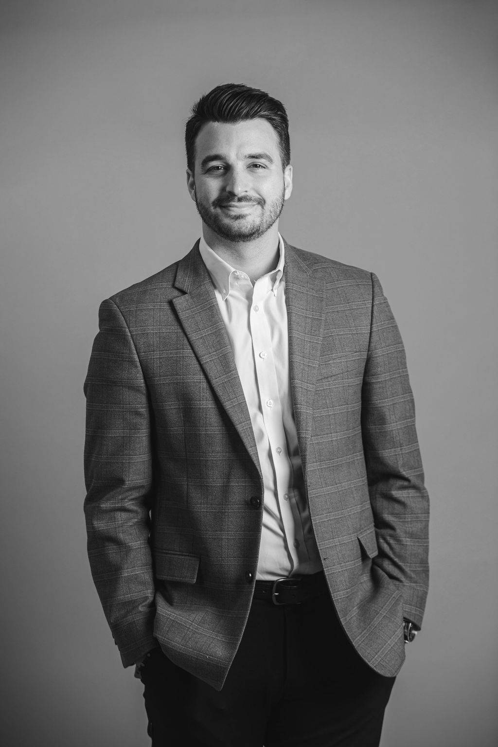 A man in a gray blazer and white shirt stands against a plain background, smiling with hands in pockets.