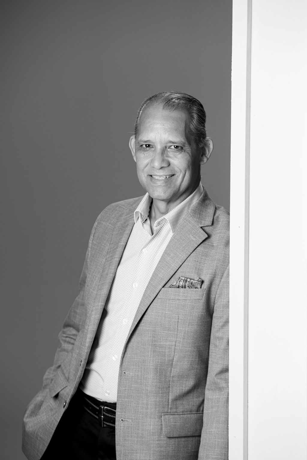 Smiling man in a light blazer leaning against a white wall, in a black-and-white portrait