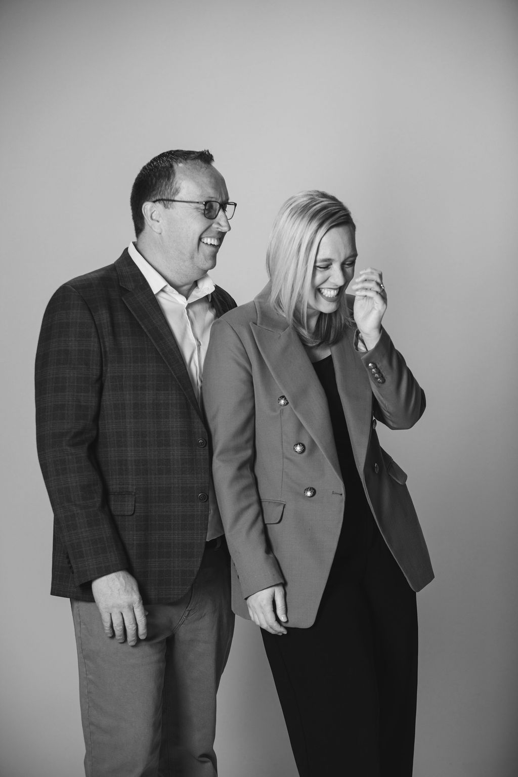 Two people standing together, smiling and looking down against a plain backdrop.
