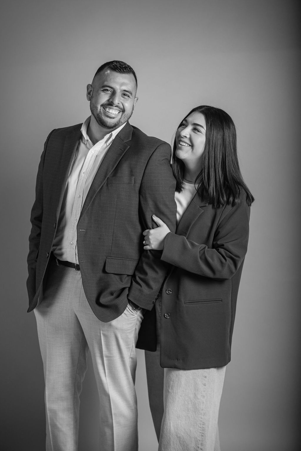 Two smiling people in suits standing together, one leaning on the other against a plain backdrop.