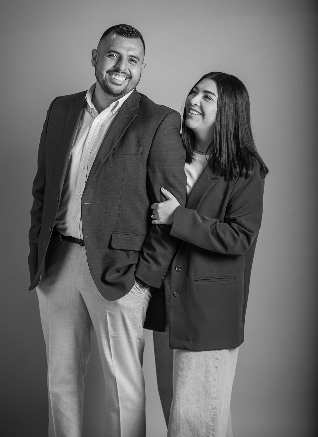 Two smiling people in blazers standing together against a plain backdrop