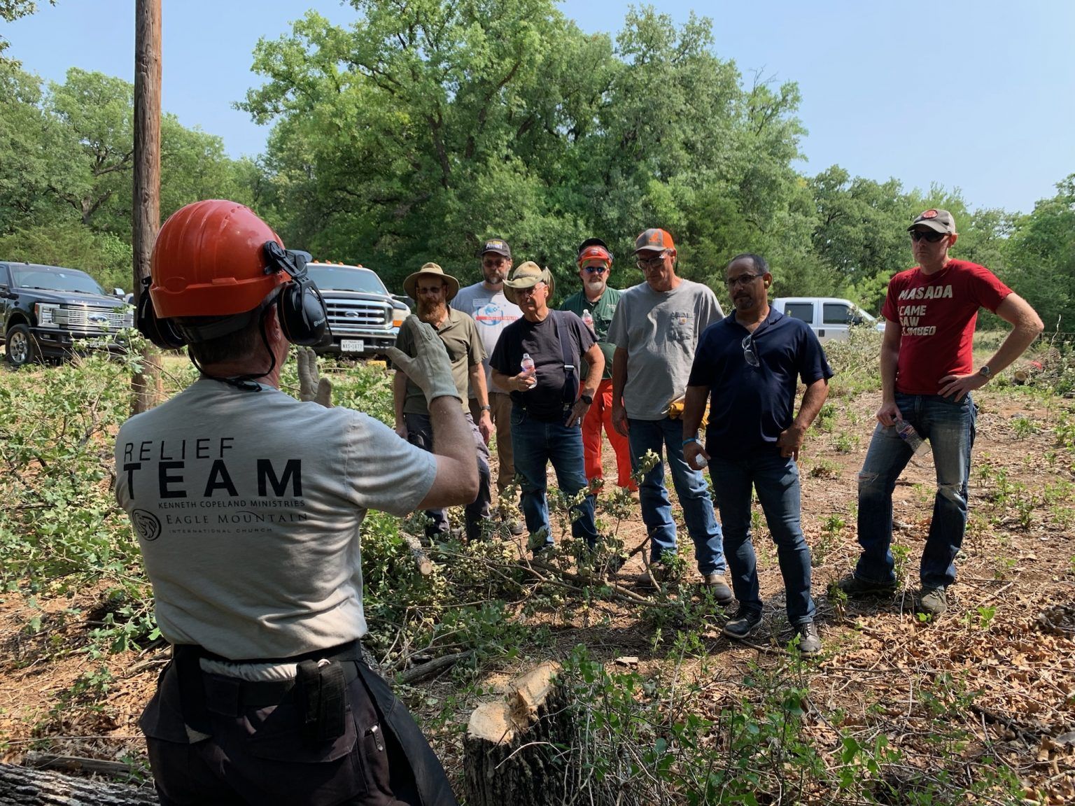 Team members in hard hats gather outdoors near trees for a site briefing while one person speaks.