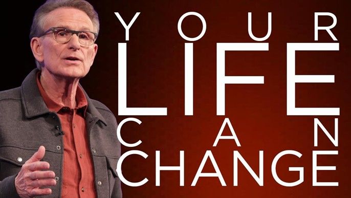 Man speaking on stage beside large text reading “YOUR LIFE A CHANGING”