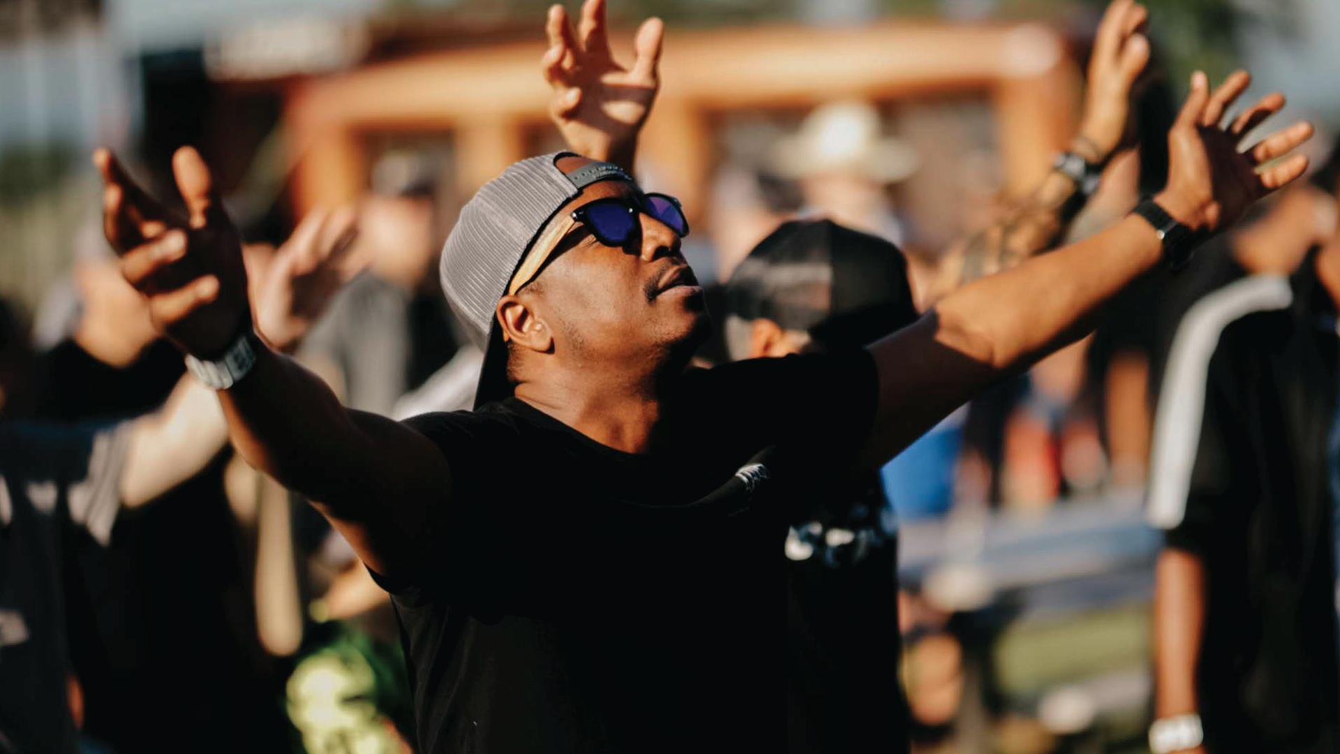 Person in a gray beanie and sunglasses raises arms amid a cheering crowd outdoors