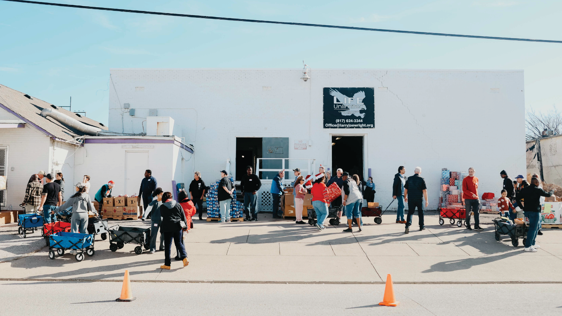 People gathered outside a white building, waiting in line near tables and orange cones.
