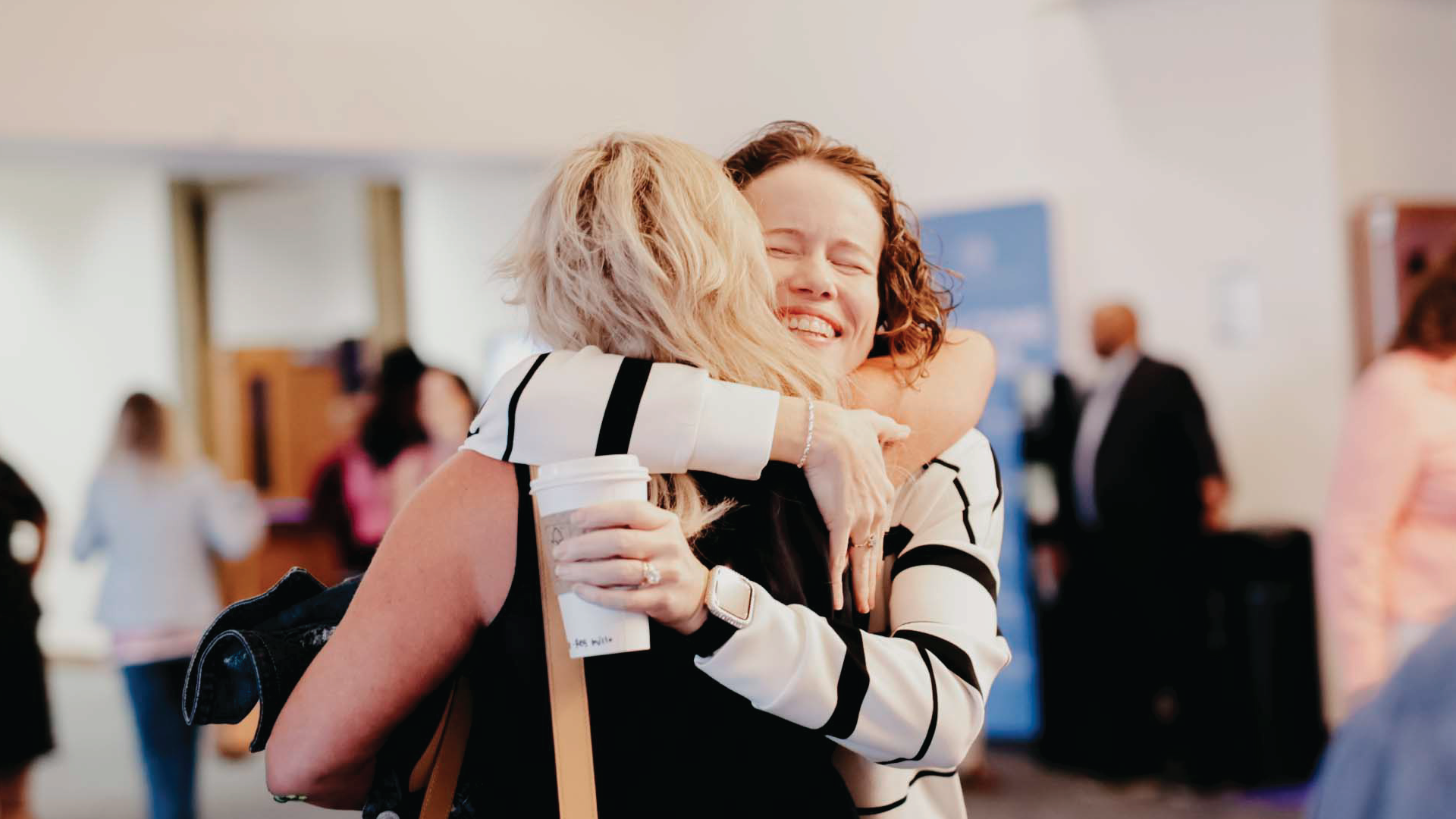 Two people hugging at a social event, one holding a coffee cup, with guests blurred in the background.