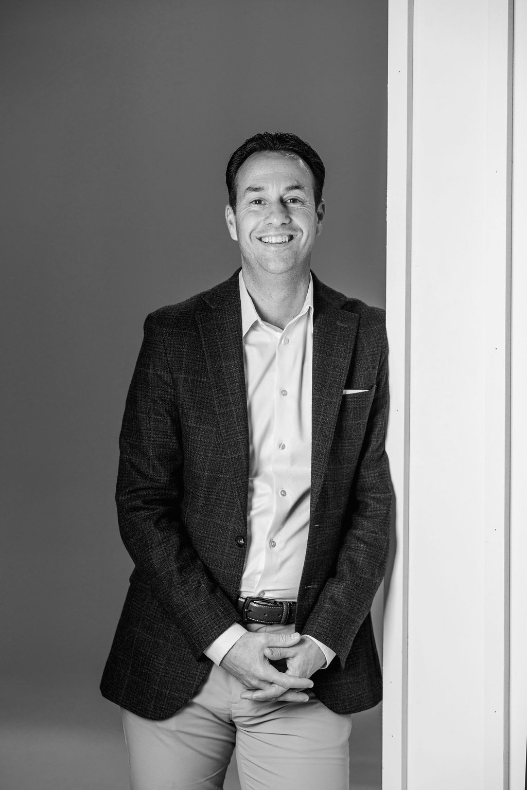 Smiling person in a dark blazer and light shirt leaning against a wall indoors