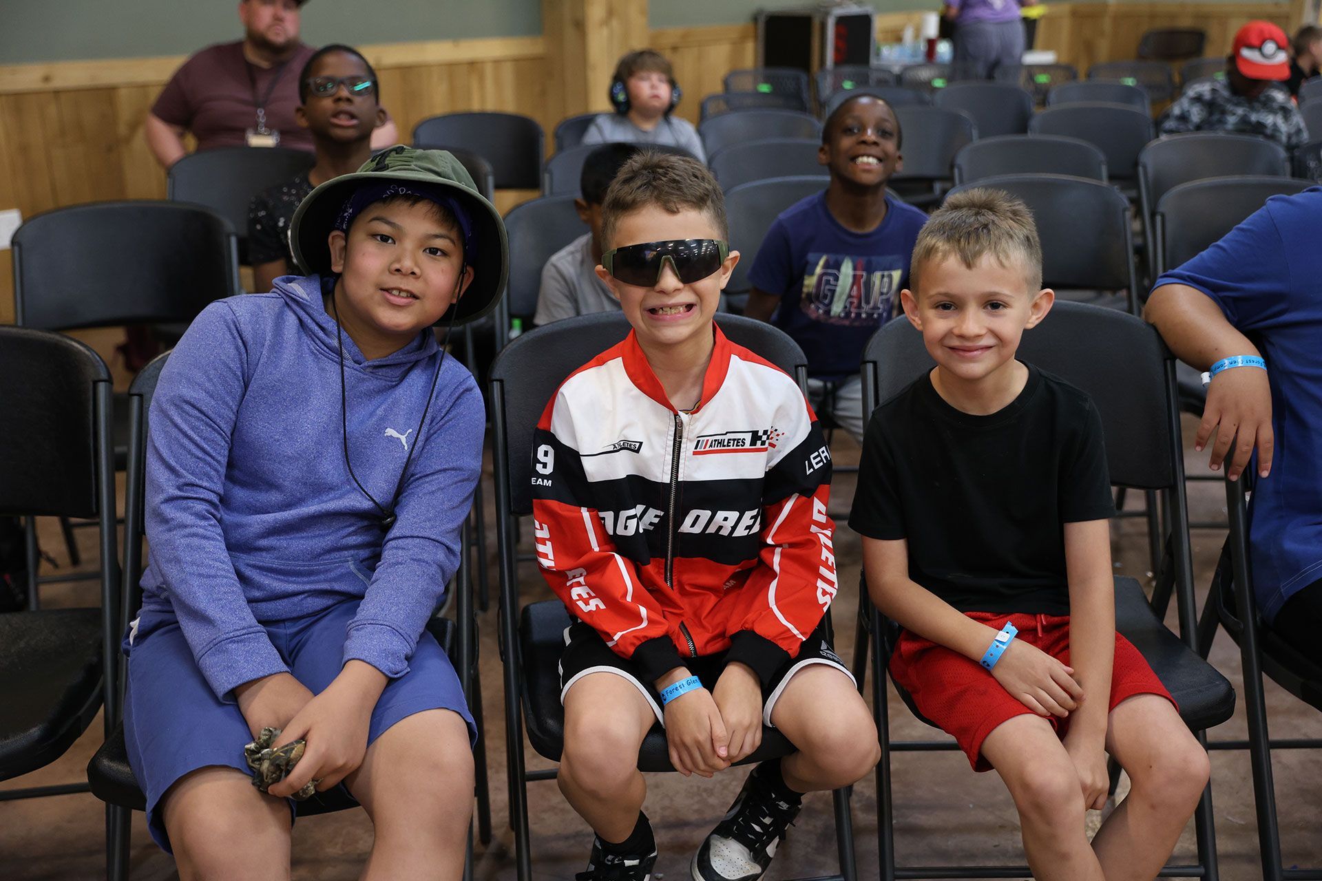 Three children seated in bleachers, smiling at the camera, in a gym or event venue.