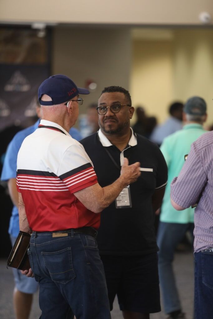 Two men talking indoors, one in a blue cap and red-striped shirt, the other in a black shirt and shorts.
