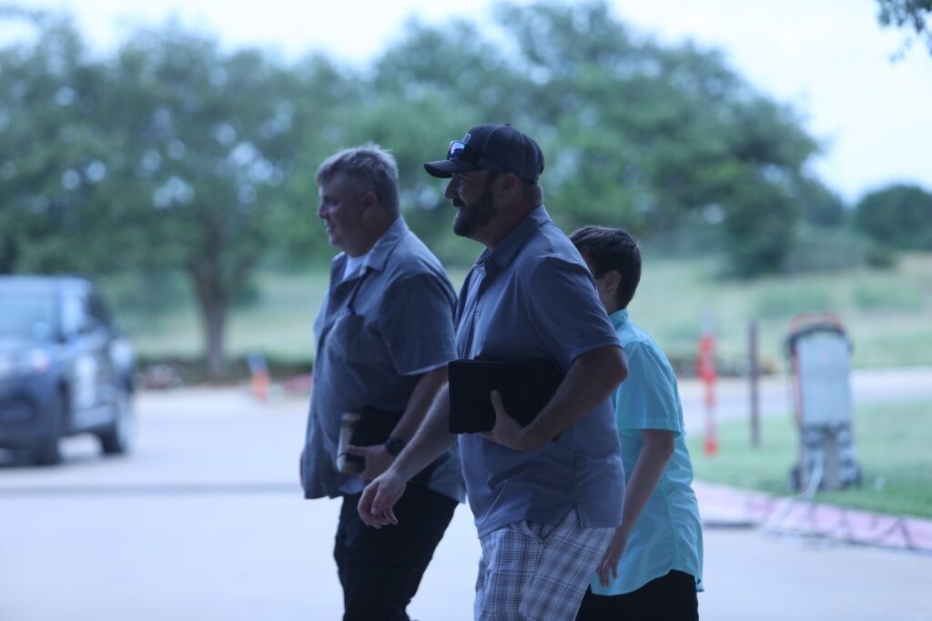 Three people walking together under a covered outdoor area, with trees and a parked vehicle in the background.