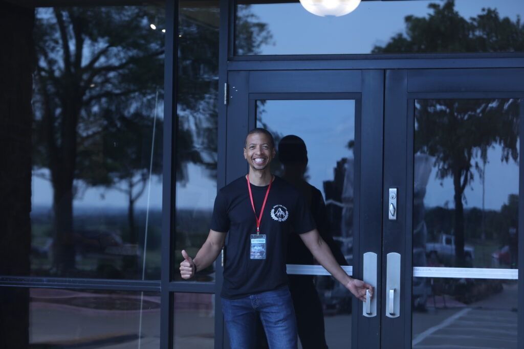 Man opening a glass door outside a building at dusk, wearing a black shirt and lanyard