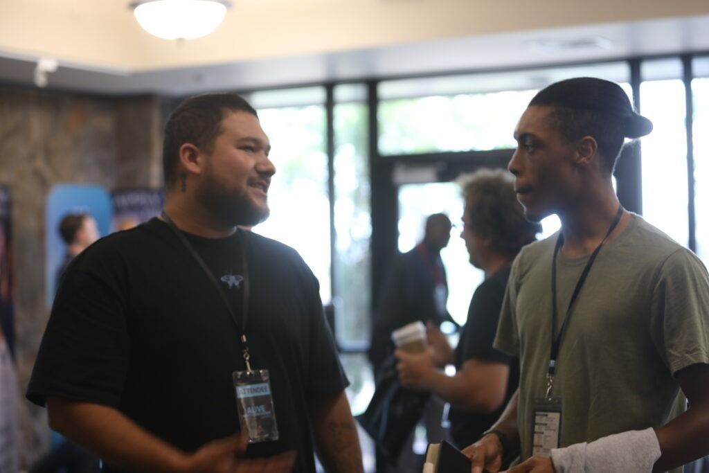 Two people talking at a conference, both wearing lanyards, in a bright lobby.