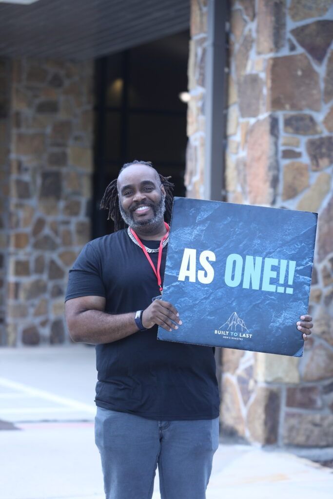 Person holding a sign that reads “AS ONE!” in front of a stone building.
