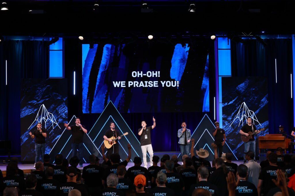 Worship band on stage under blue lights, with screen text reading “OH-OH! WE PRAISE YOU!”