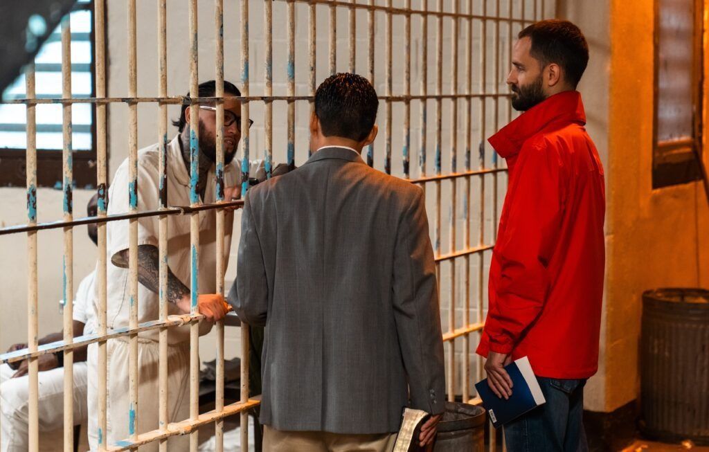 Two people stand at a metal gate speaking with a man in a white coat inside a room