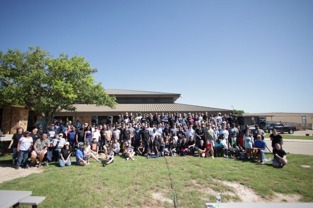 Large group posing outdoors on a sunny day in front of a building with a tree and grassy foreground