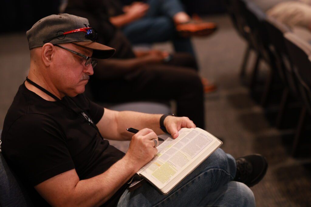 Man in a black shirt and cap writing in a notebook while seated in an auditorium