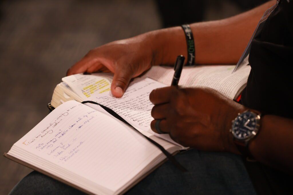 Hands writing in a notebook with a pen, with another open book beneath on a desk.