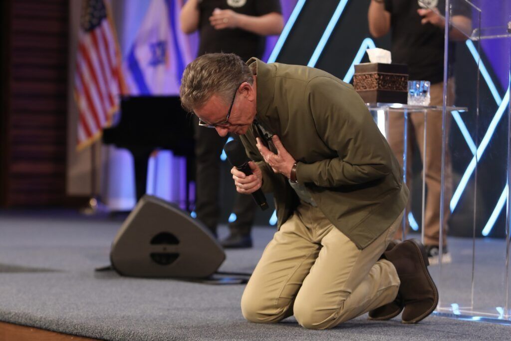 Man kneels on a stage, praying with head bowed, holding a microphone near his chest.