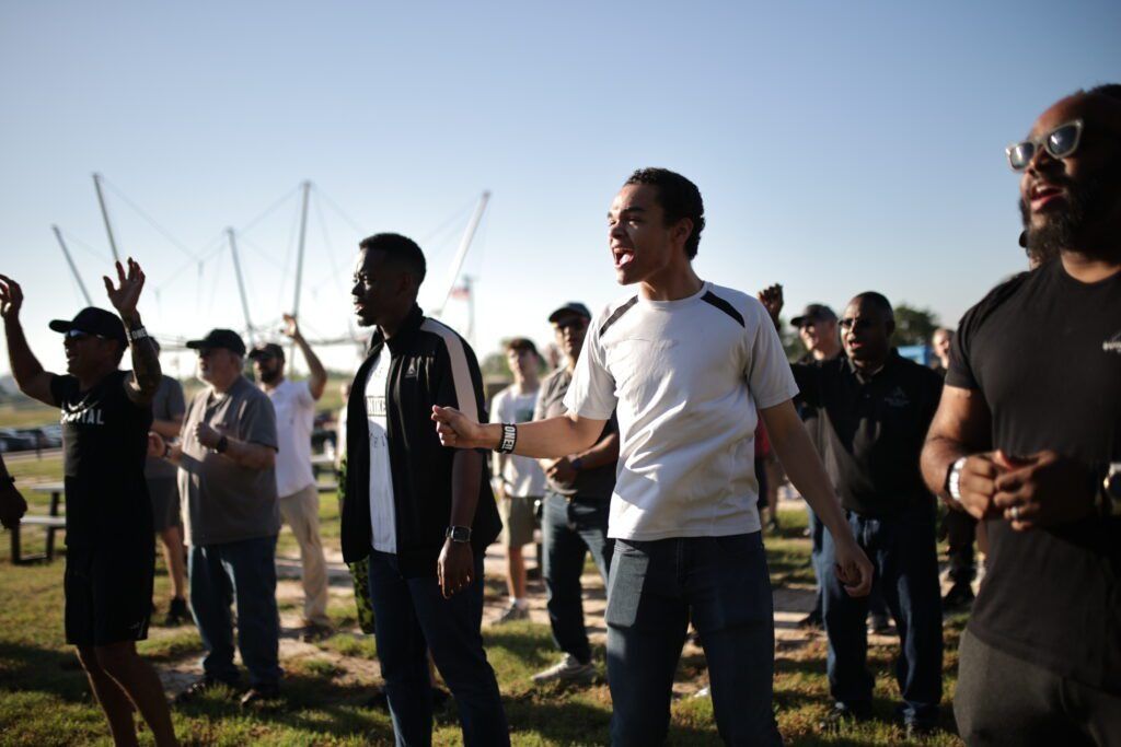 Group of people standing outdoors in a sunny field, some raising hands, with kites in the sky
