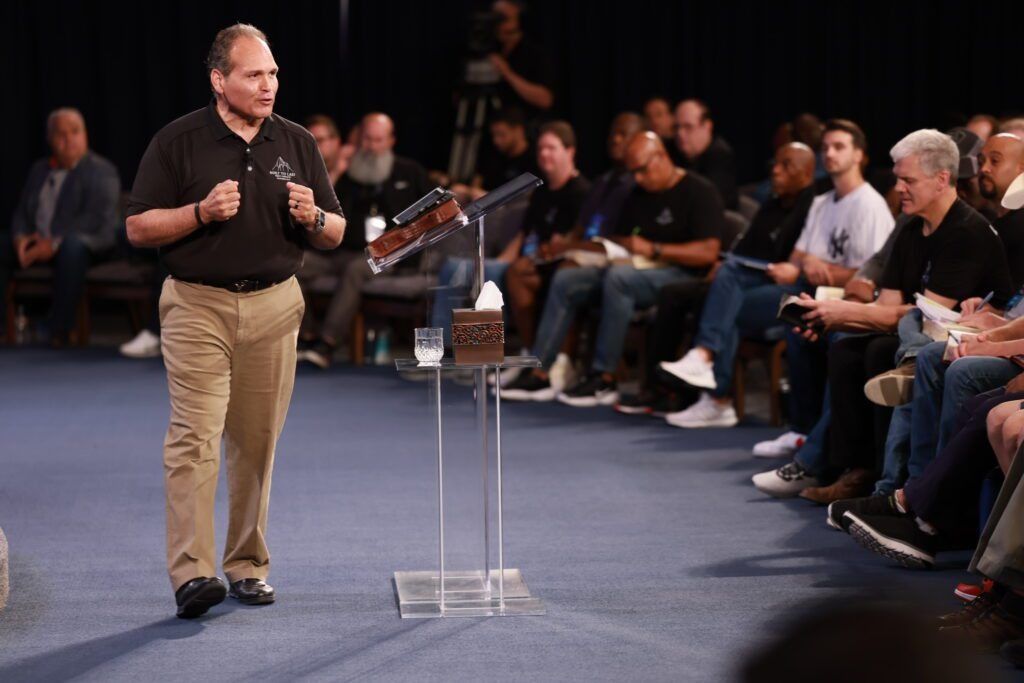 Man speaking on stage beside a clear lectern, facing an audience seated in rows