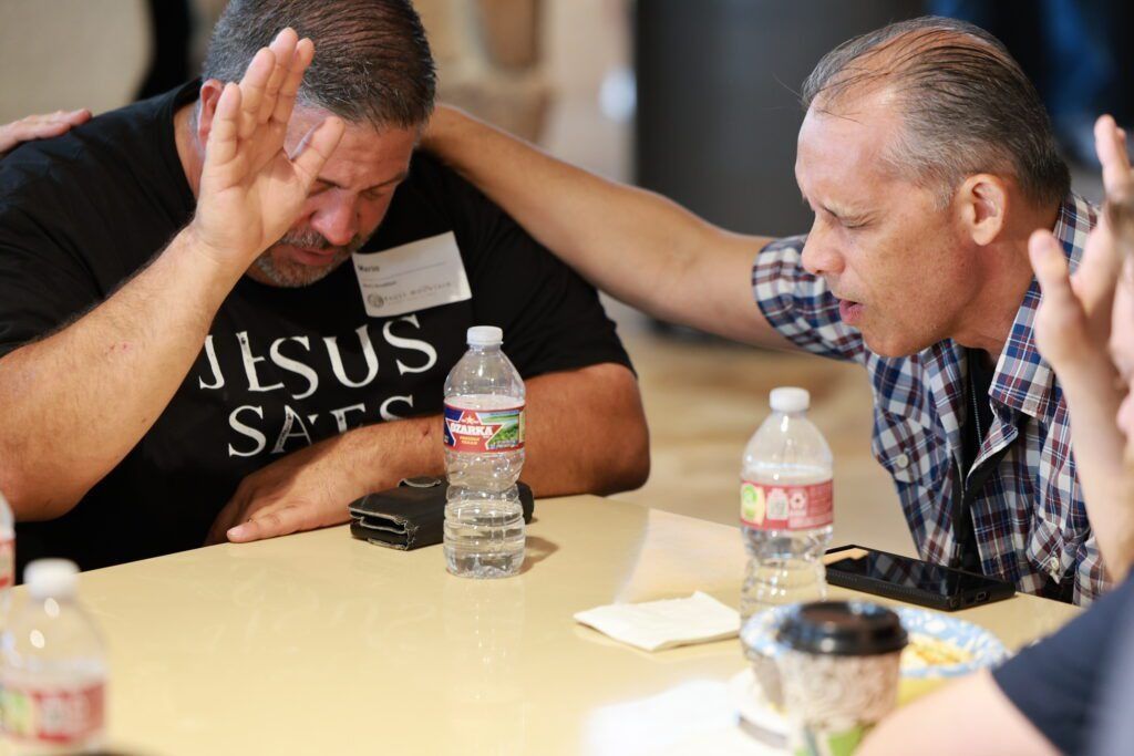 Two men at a table in prayer or discussion, heads bowed and hands raised, with water bottles nearby.