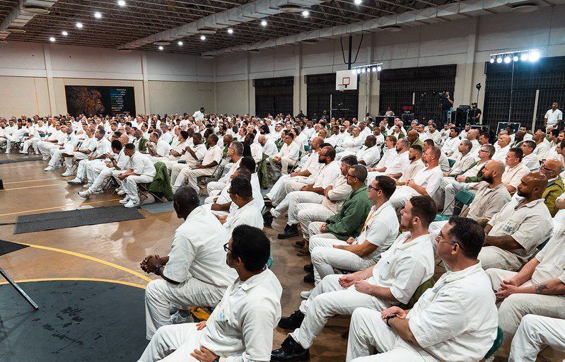 Large seated audience in a conference hall, many in white clothing, listening attentively to a speaker.