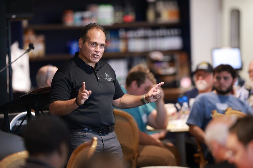 Speaker addressing a seated audience in a casual indoor meeting, gesturing with both hands.