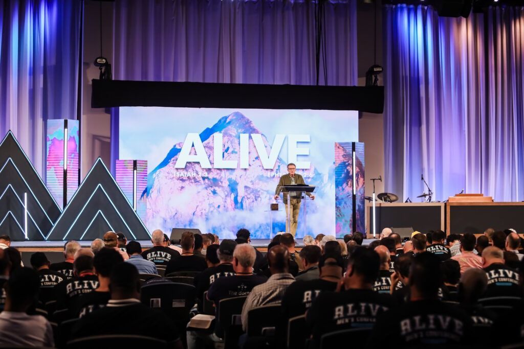 Conference audience watching a speaker onstage beneath a large “ALIVE” screen with purple lighting