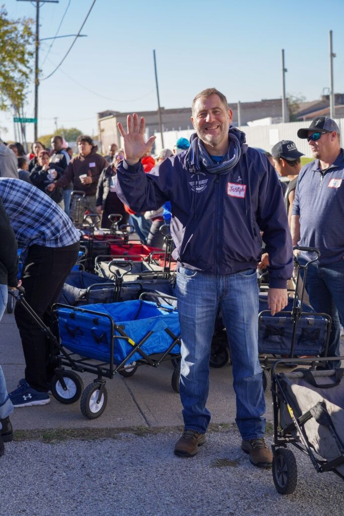 Man smiling and waving at an outdoor event with carts and bags in the background
