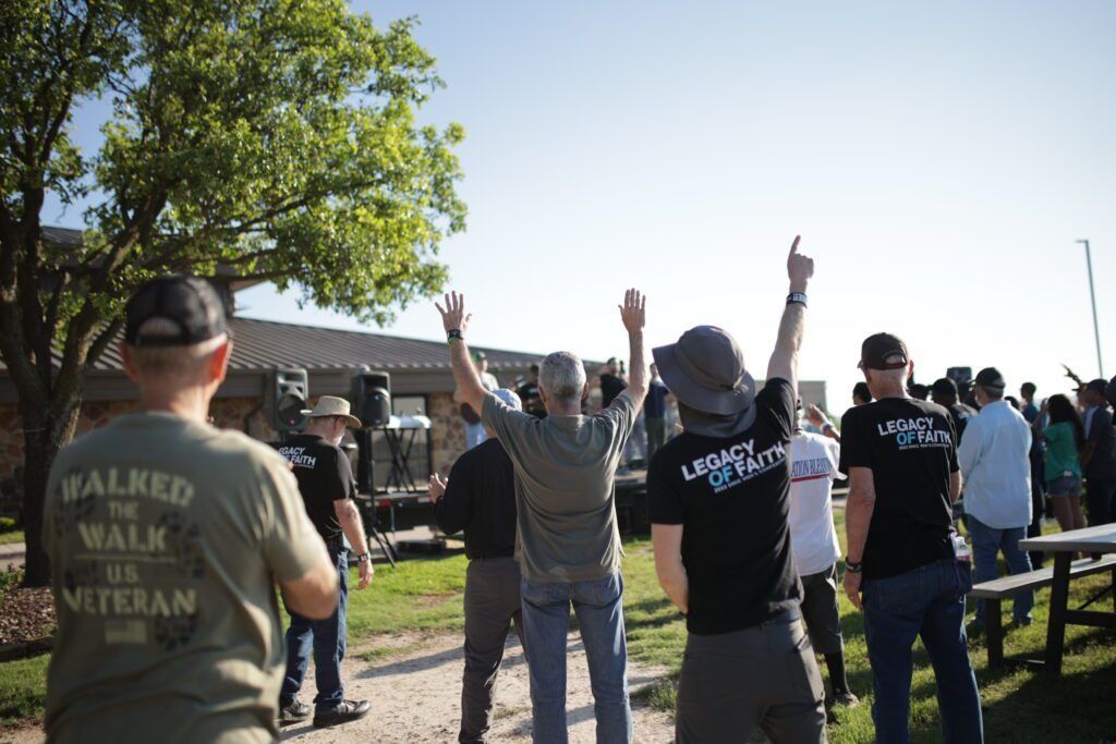 People with raised hands during a walk event in a sunny park, with a tree and open field behind them